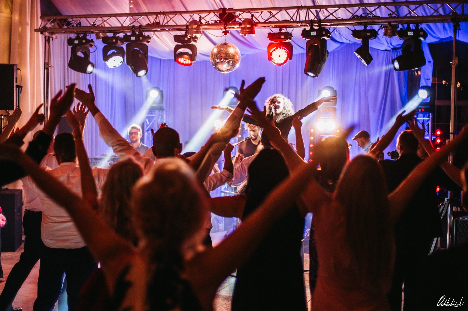 Zespół muzyczny na scenie podczas występu, wokalistka z rozłożonymi rękami, publiczność z uniesionymi rękami, oświetlenie sceniczne i kula dyskotekowa.