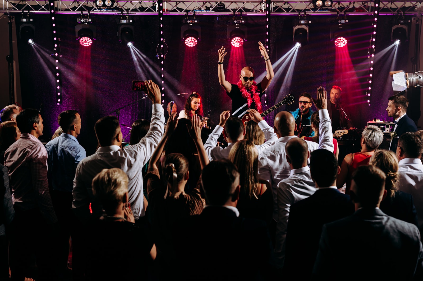 Występujący na scenie wokalista w okularach przeciwsłonecznych i różowym boa, z gitarą elektryczną, w tle oświetlenie sceniczne i publiczność z podniesionymi rękami.