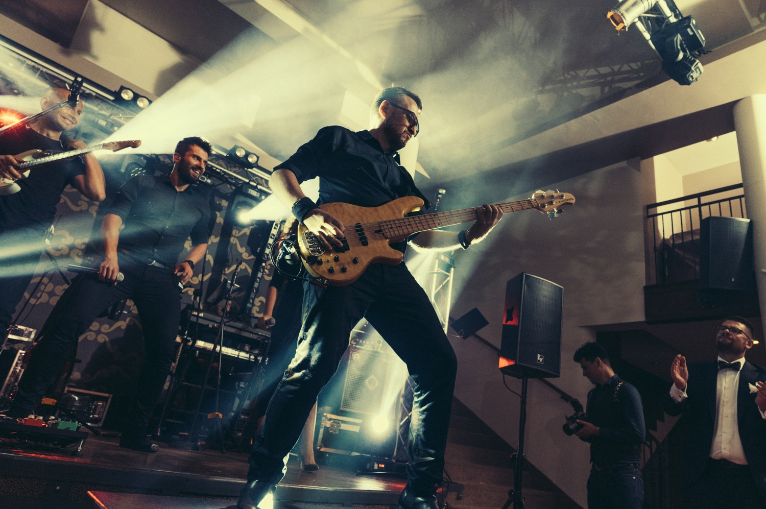 Basista w okularach grający na basie podczas występu na scenie oświetlonej reflektorami, obok wokalista z mikrofonem i gitarzysta.