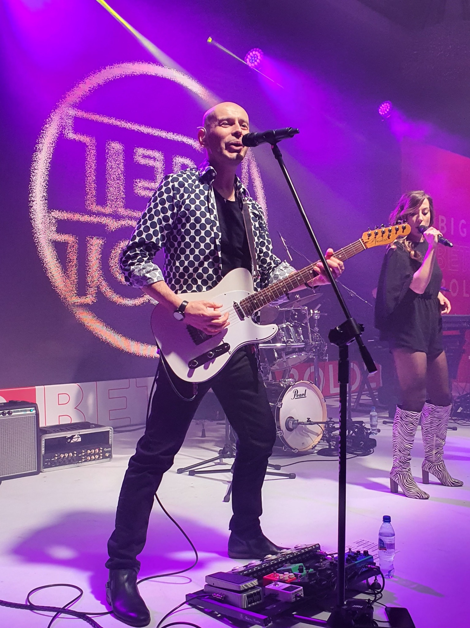 Wokalista z gitarą w koszuli w grochy, w tle wokalistka w kozakach w zebrę, na scenie perkusja i efekty gitarowe, oświetlenie sceniczne w odcieniach fioletu.