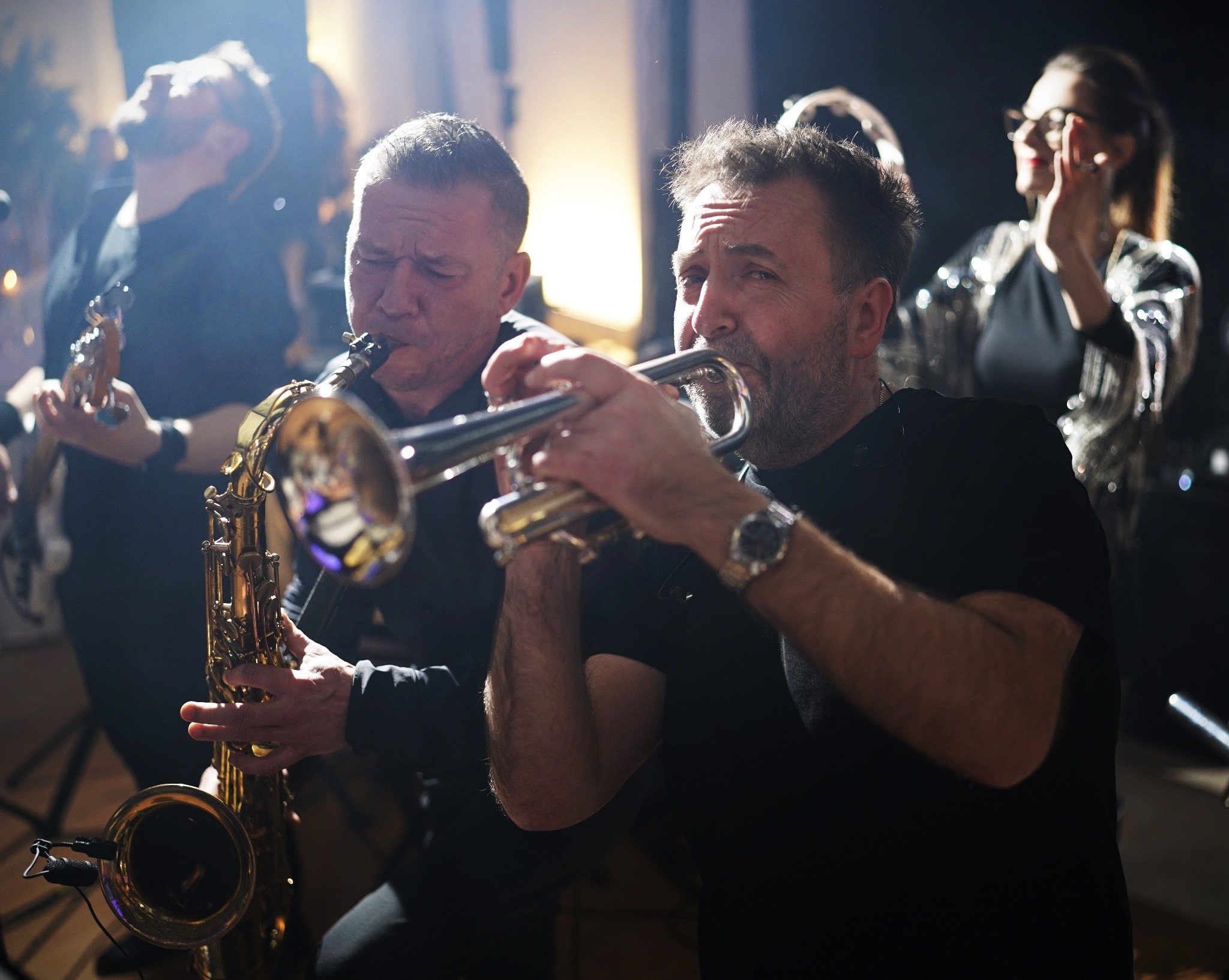 Dwóch muzyków grających na saksofonie i trąbce podczas występu na scenie, w tle pozostali członkowie zespołu i publiczność.