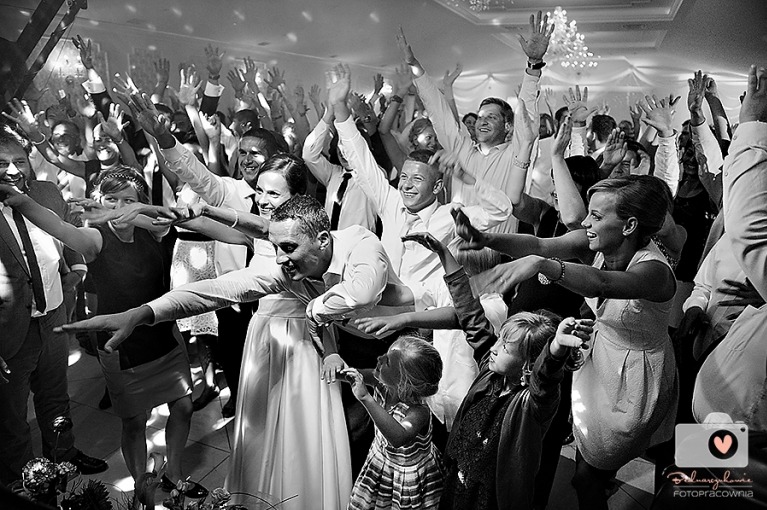 Czarno-białe zdjęcie rozbawionego tłumu gości weselnych tańczących na parkiecie z podniesionymi rękami, w tle dekoracje sali.