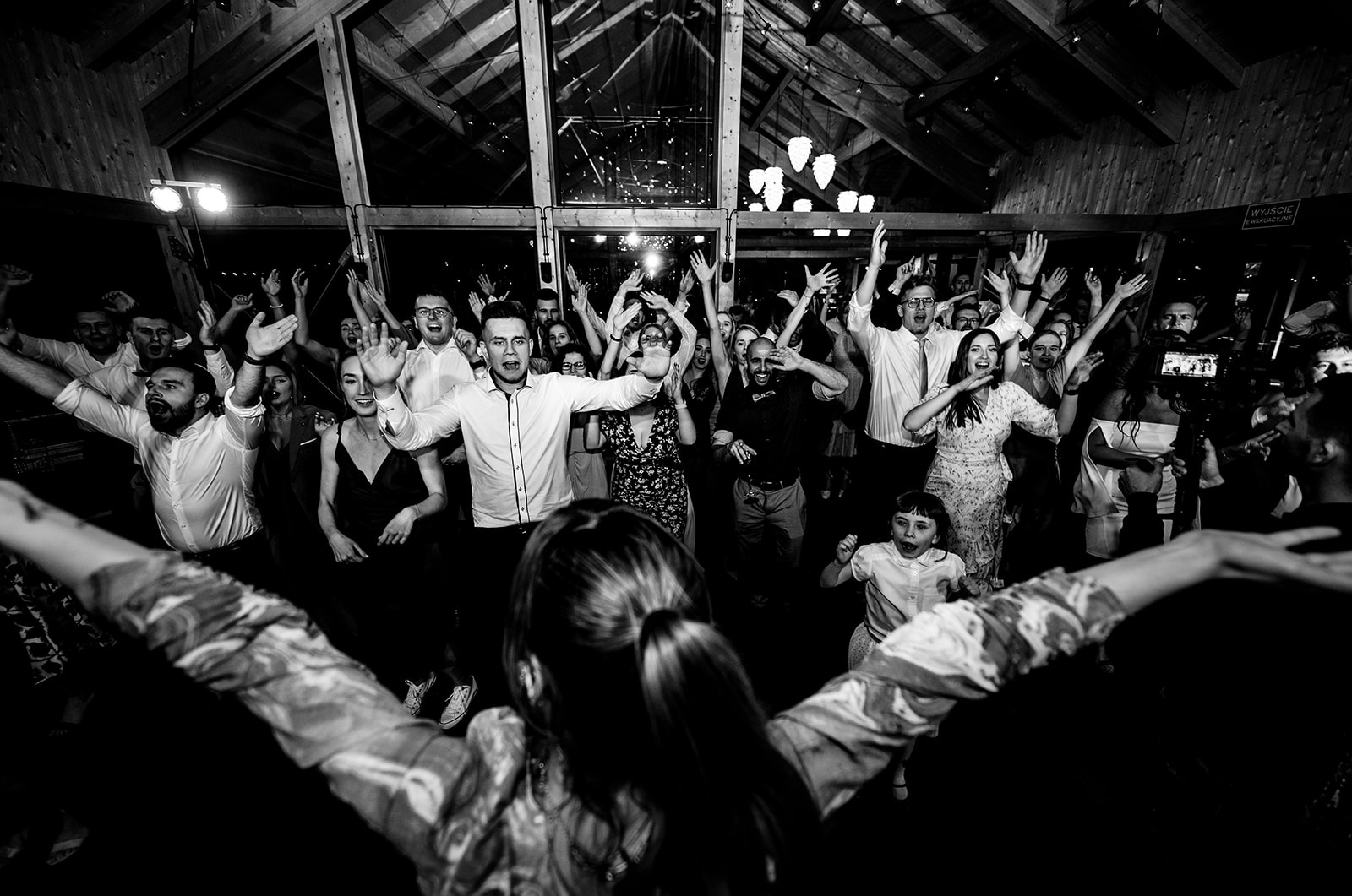 Czarno-białe zdjęcie rozbawionego tłumu z podniesionymi rękami, widzianego z perspektywy osoby z tyłu, podczas imprezy w drewnianym wnętrzu z oknami i dekoracyjnym oświetleniem.