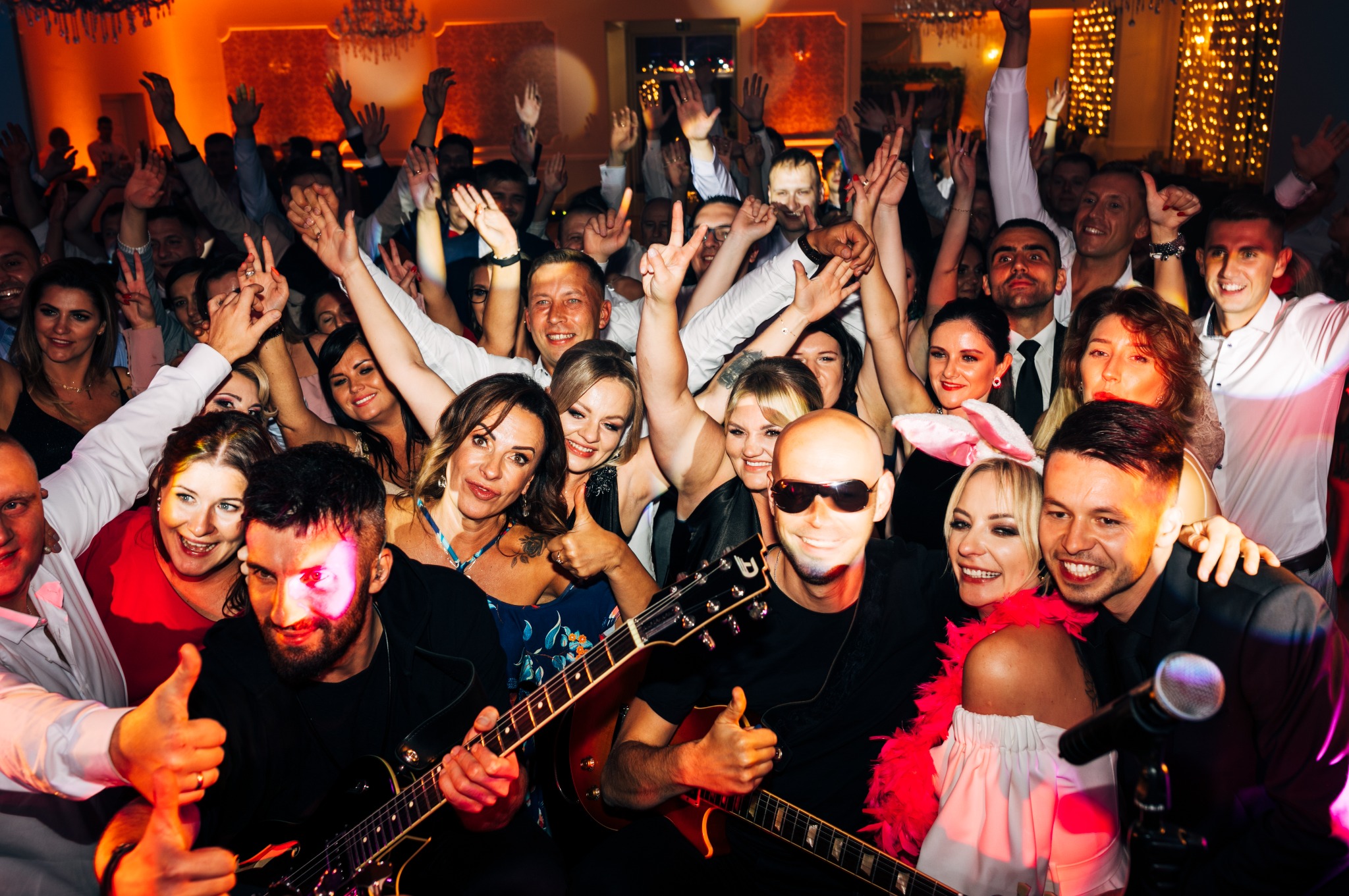 Rozentuzjazmowany tłum gości weselnych z podniesionymi rękami, w pierwszym planie gitarzyści z uniesionymi kciukami w górę, w tle dekoracyjne oświetlenie sali.