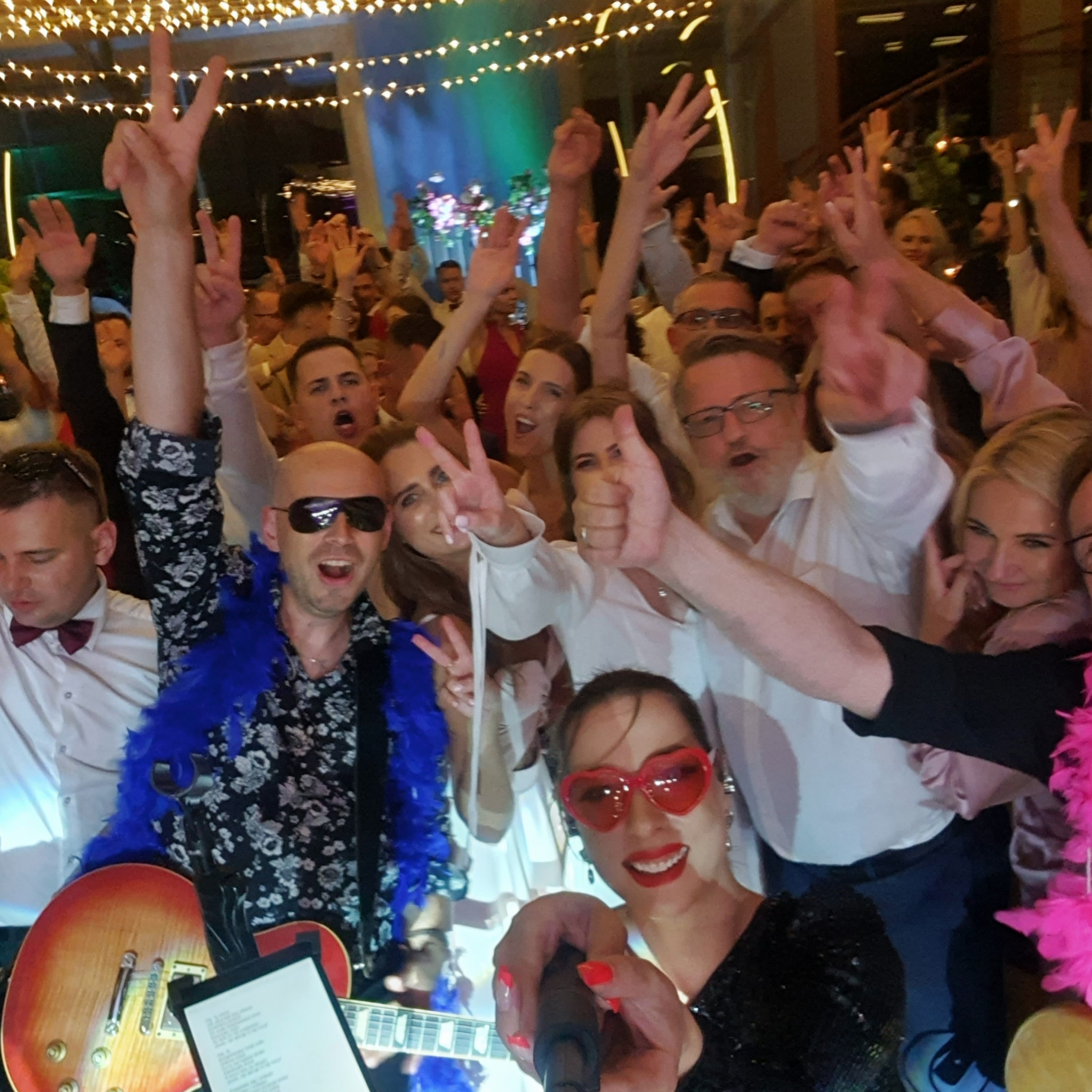 Selfie z wokalistką w czerwonych okularach w kształcie serc i gitarzystą w niebieskim boa na tle rozbawionego tłumu podczas imprezy.