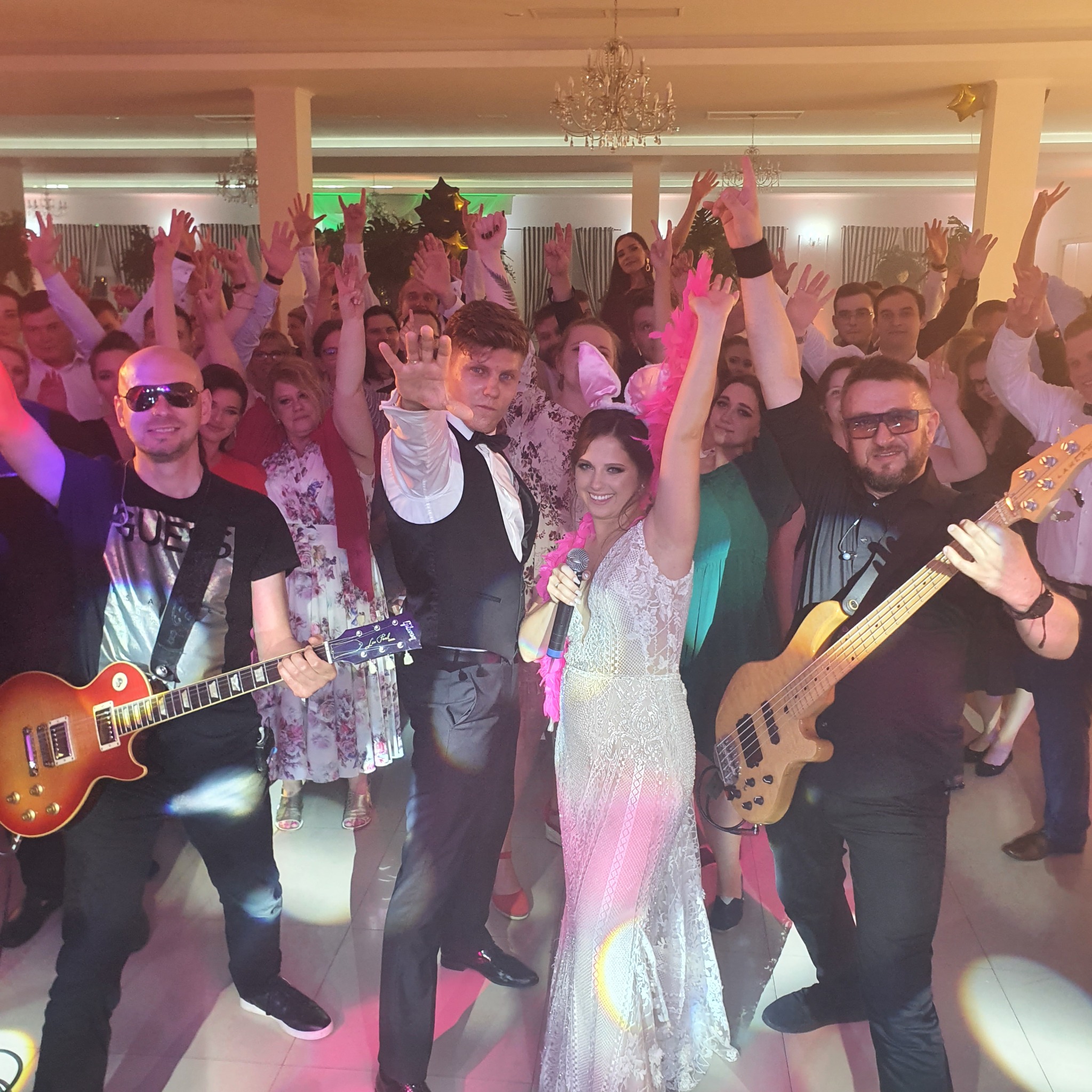 Wokalistka w koronkowej sukni z mikrofonem i różowym boa z pierza, obok niej mężczyzna w smokingu, dwóch gitarzystów z gitarami elektrycznymi oraz rozbawiony tłum gości weselnych z podniesionymi...