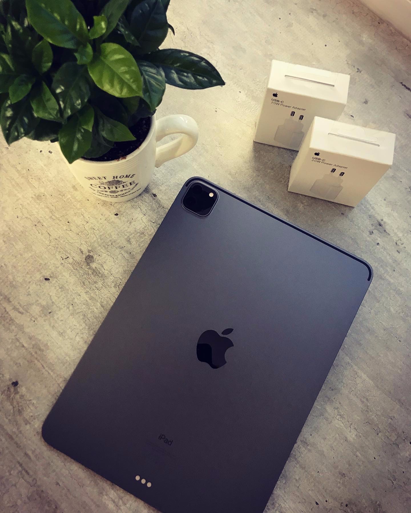 Nowy, grafitowy tablet z logo nadgryzionego jabłka leżący na szarej powierzchni, obok doniczki z zieloną rośliną w białym kubku z napisem 'Sweet Home Coffee' oraz dwóch pudełek z zasilaczami USB-C...