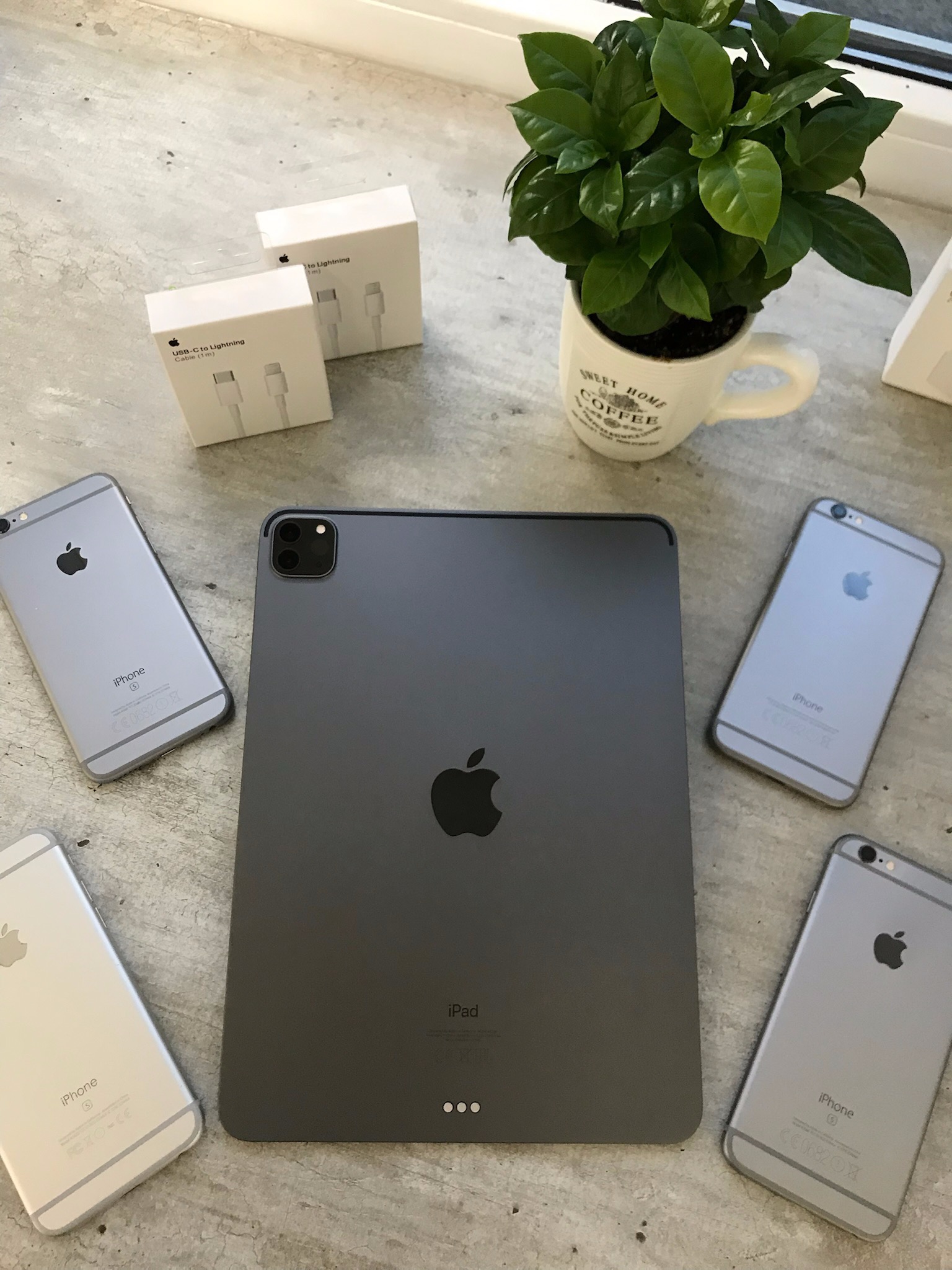 Centralnie umieszczony ciemnoszary iPad Pro otoczony czterema iPhone'ami 6S, z pudełkami kabli USB-C do Lightning i małą rośliną doniczkową w tle, na szarej powierzchni.