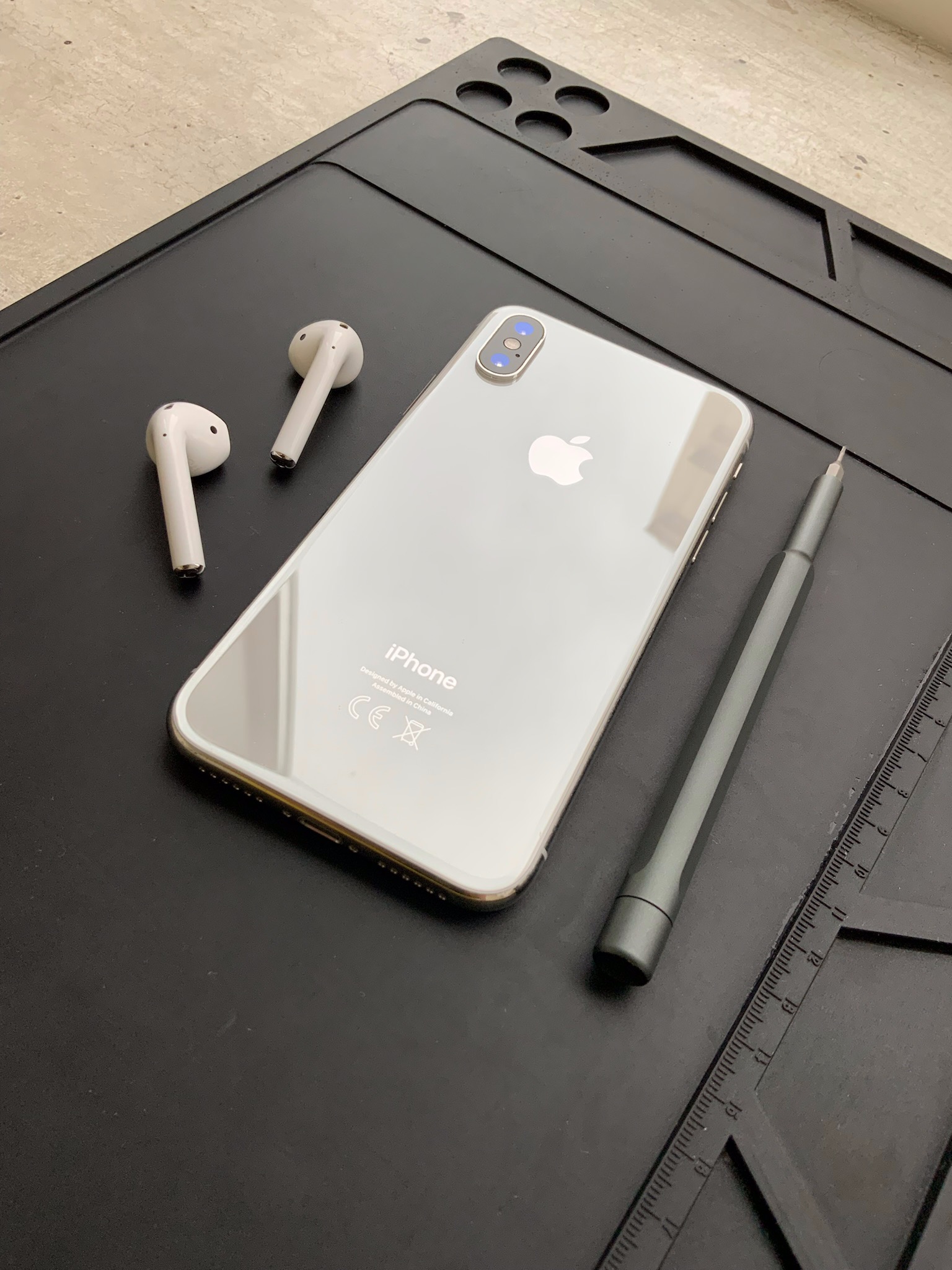 Srebrny iPhone X, białe słuchawki AirPods oraz precyzyjny śrubokręt leżą na czarnej, antypoślizgowej macie serwisowej z podziałką.