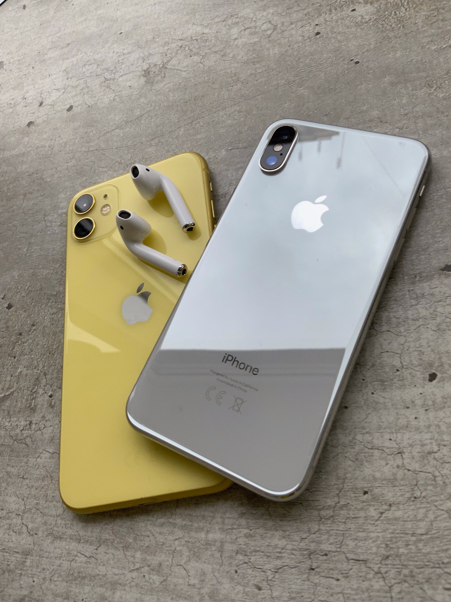 Dwa smartfony iPhone, jeden żółty i jeden srebrny, leżące na szarej, betonowej powierzchni, z parą słuchawek AirPods na żółtym telefonie.