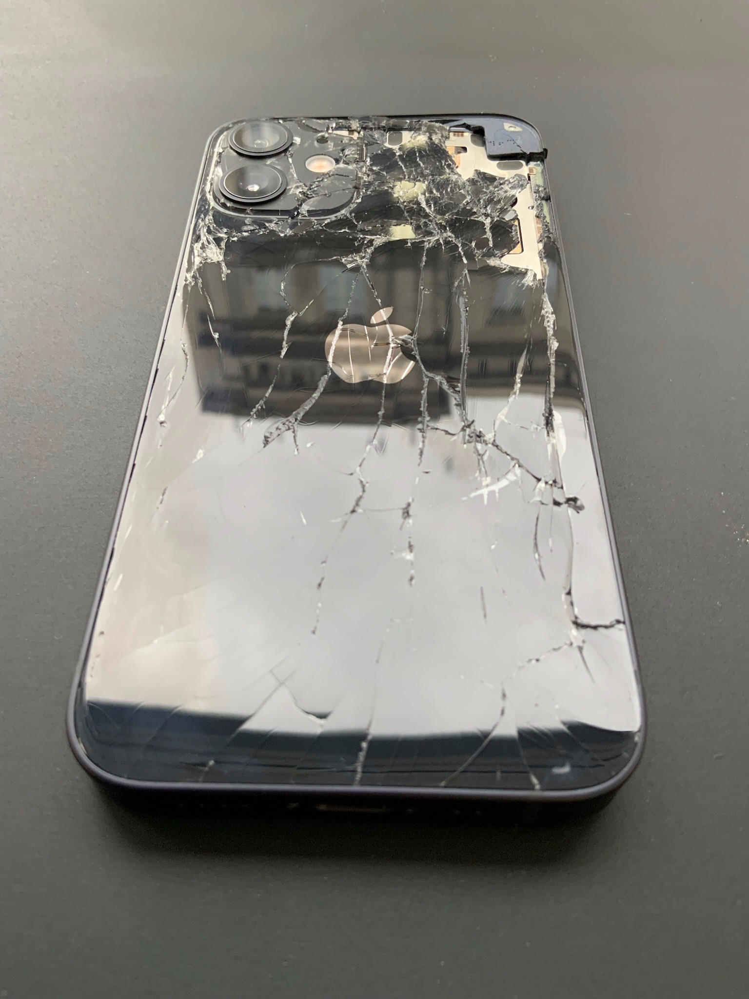 Smartfon Apple iPhone z mocno potłuczoną szklaną obudową, widoczne pęknięcia i ubytki szkła, uszkodzone narożniki i odsłonięte wnętrze obok aparatu.