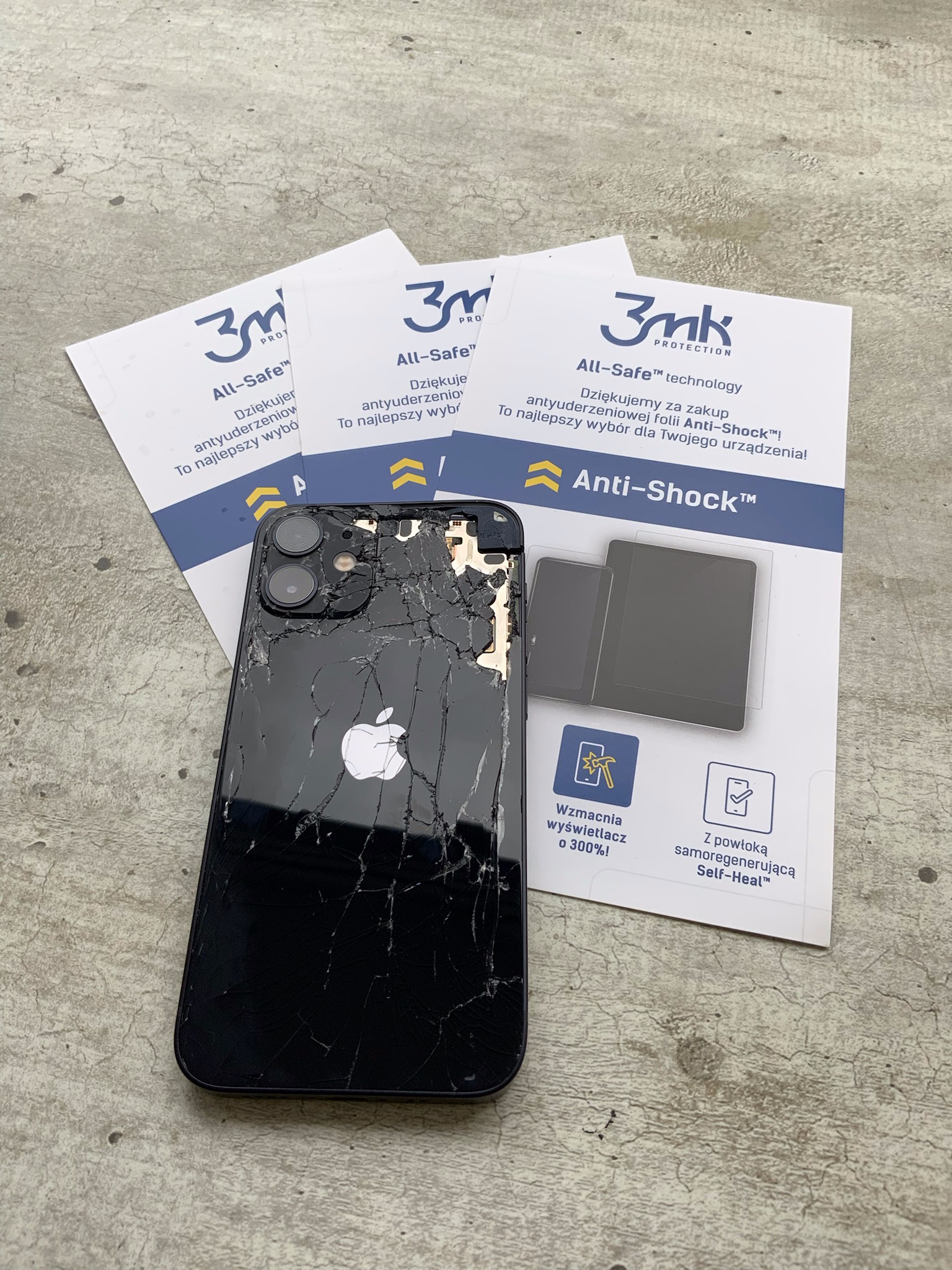 Rozbity czarny smartfon leżący obok trzech opakowań folii ochronnej 3mk All-Safe na szarym, betonowym tle. Ekran i tylna obudowa telefonu są popękane w pajęczynę.