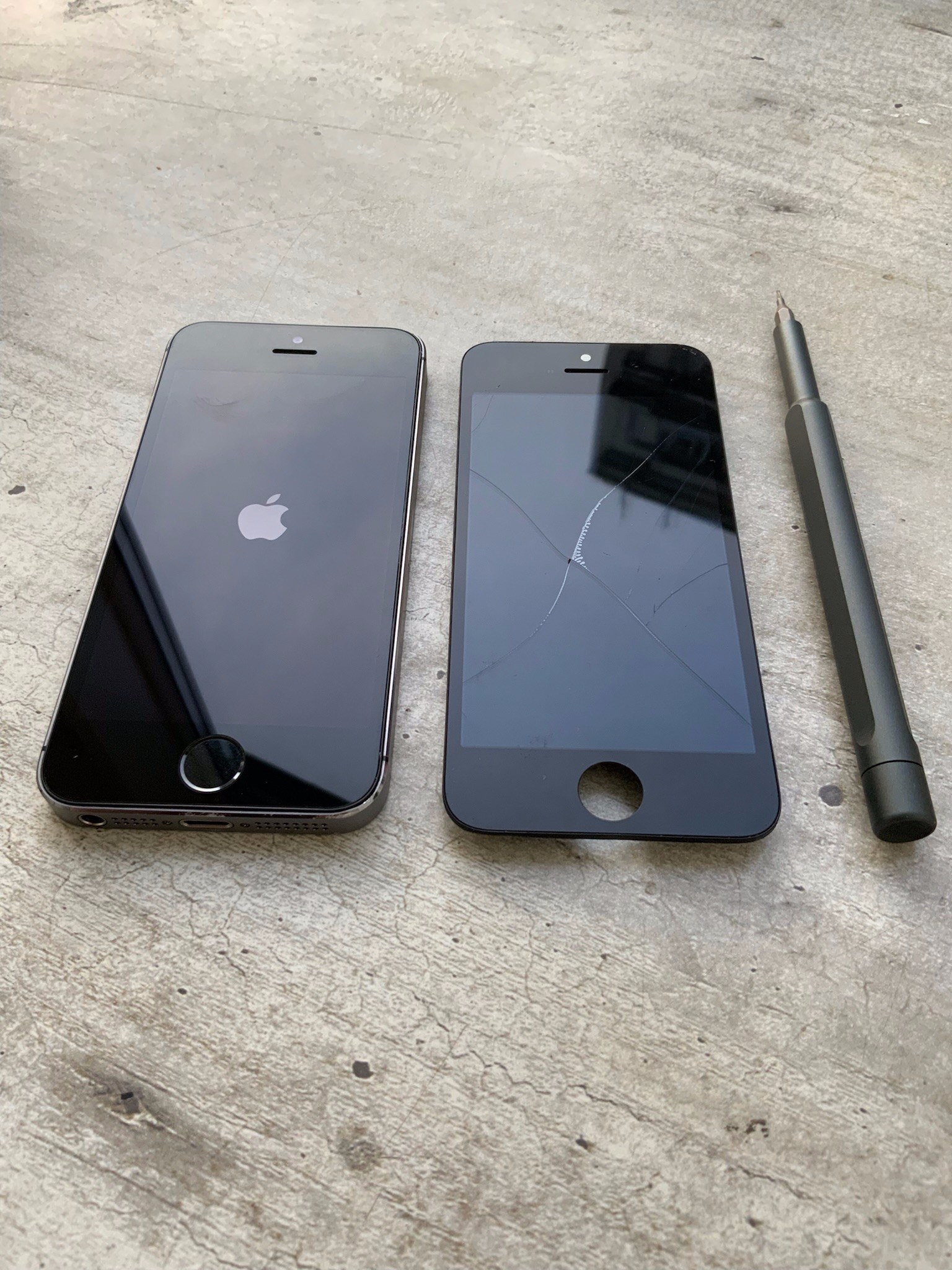 Dwa czarne telefony iPhone leżące obok siebie na szarym betonowym tle, jeden z włączonym ekranem z logo Apple, drugi z pękniętym ekranem, obok leży szary śrubokręt.