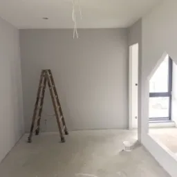Wnętrze pokoju po malowaniu na szaro, widoczna drewniana drabina, okno w kształcie domku i puszka z farbą na podłodze.