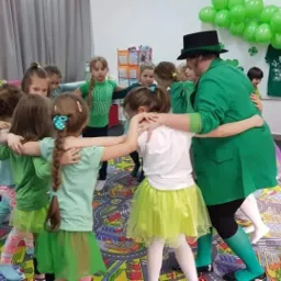 Grupa dzieci i animator w zielonym stroju tworzą krąg podczas zabawy z okazji Dnia Świętego Patryka, widoczne zielone balony i dekoracje tematyczne.