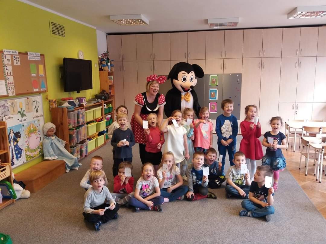 Grupa roześmianych dzieci pozuje do zdjęcia z animatorką w czerwonej sukience w grochy i postacią w kostiumie Myszki Miki w przedszkolu.