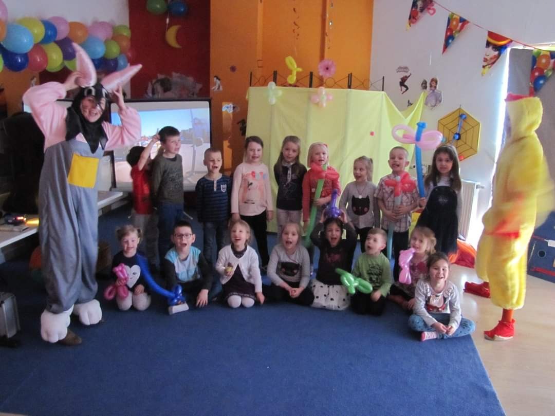 Grupa dzieci pozuje do zdjęcia z animatorem przebranym za królika i osobą w kostiumie kurczaka na tle dekoracji urodzinowych.