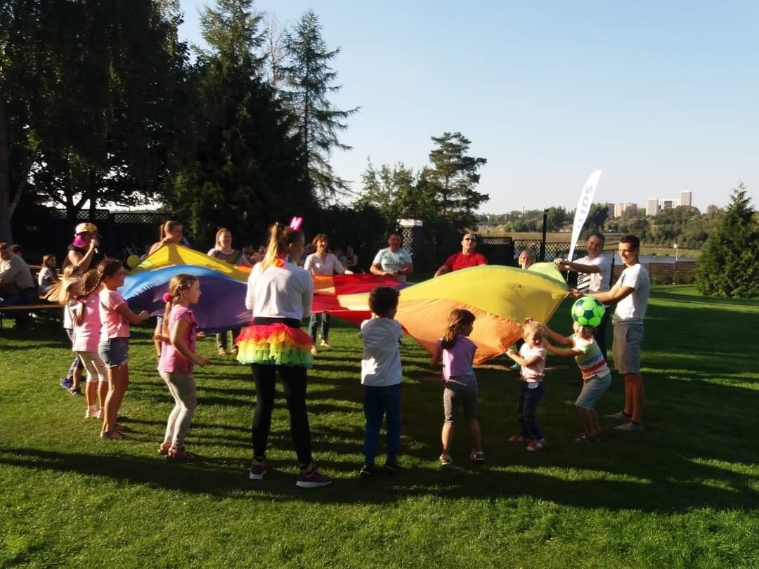 Grupa dzieci i dorosłych bawi się kolorową chustą animacyjną na zielonej trawie w plenerze; w tle drzewa i budynki.