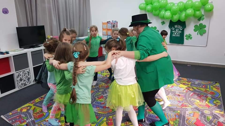 Grupa dzieci i animator w zielonym stroju tworzą krąg podczas zabawy z okazji Dnia Świętego Patryka, widoczne zielone balony i dekoracje tematyczne.