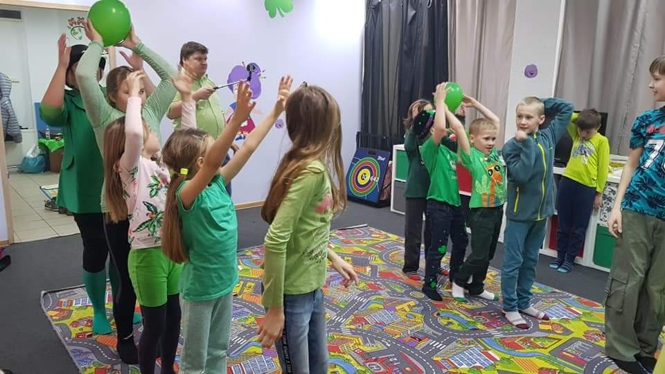 Grupa dzieci i dorosły w zielonych strojach bawią się w pomieszczeniu z kolorową podłogą, trzymając zielone balony nad głowami.