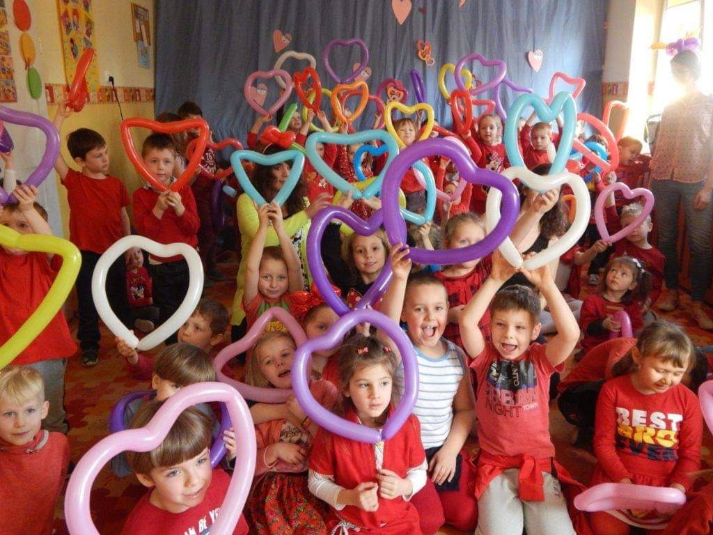 Grupa dzieci w czerwonych ubraniach trzyma balony w kształcie serc różnych kolorów podczas wydarzenia w pomieszczeniu.