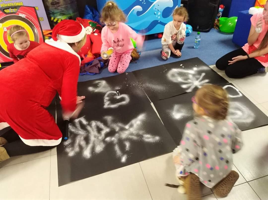 Osoba w stroju Świętego Mikołaja maluje białą farbą w sprayu wzory na czarnym papierze, wokół niej dzieci obserwują proces twórczy i trzymają w rękach rekwizyty.