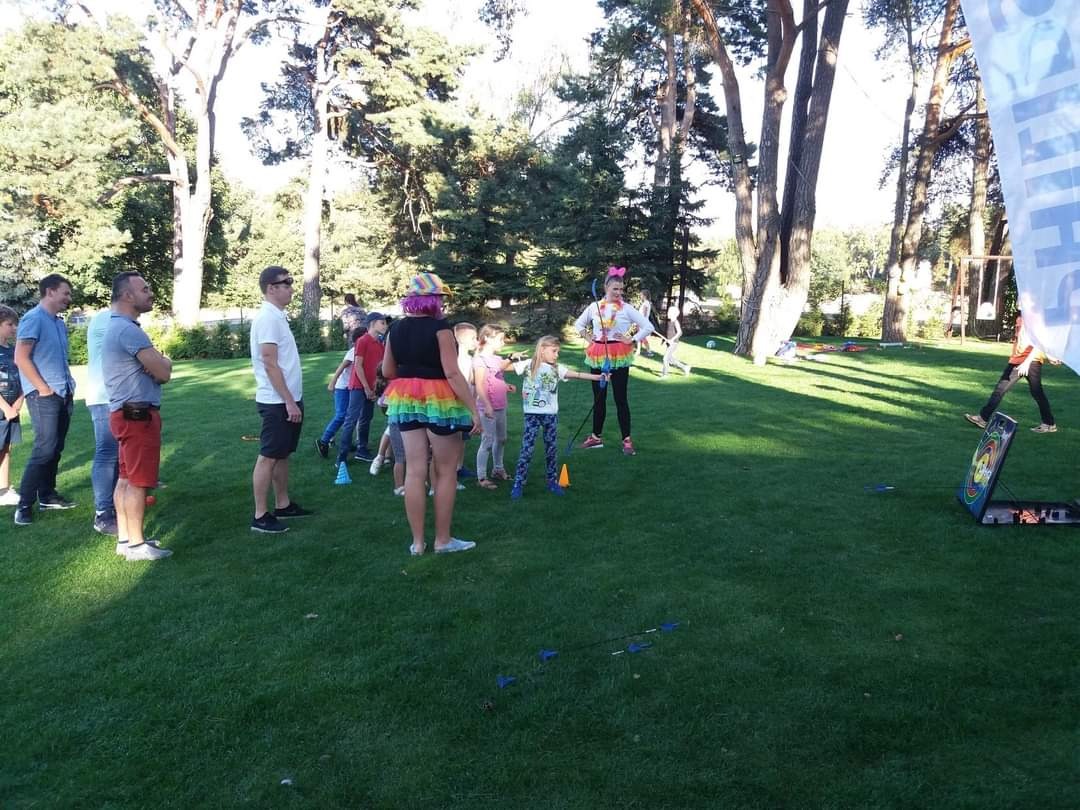 Animatorzy prowadzą zajęcia łucznicze dla dzieci na trawniku, w tle drzewa i obserwujący rodzice.