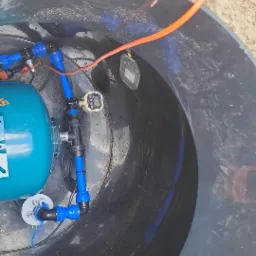 Wnętrze studni głębinowej z niebieskimi rurami, zbiornikiem hydroforowym i elektronicznym licznikiem. Widok z góry na instalację w czarnej obudowie.