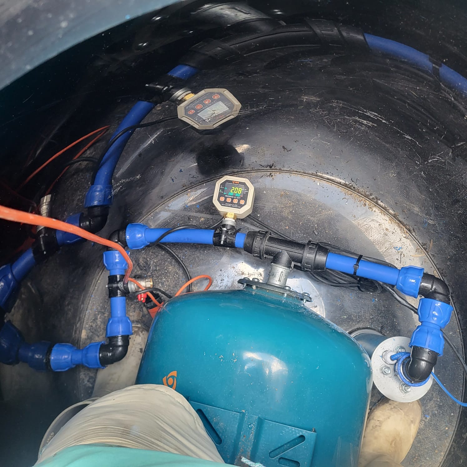 Instalacja pompy głębinowej w zbiorniku, z widocznymi niebieskimi rurami, manometrami i elementami przyłączeniowymi. Widok z góry.