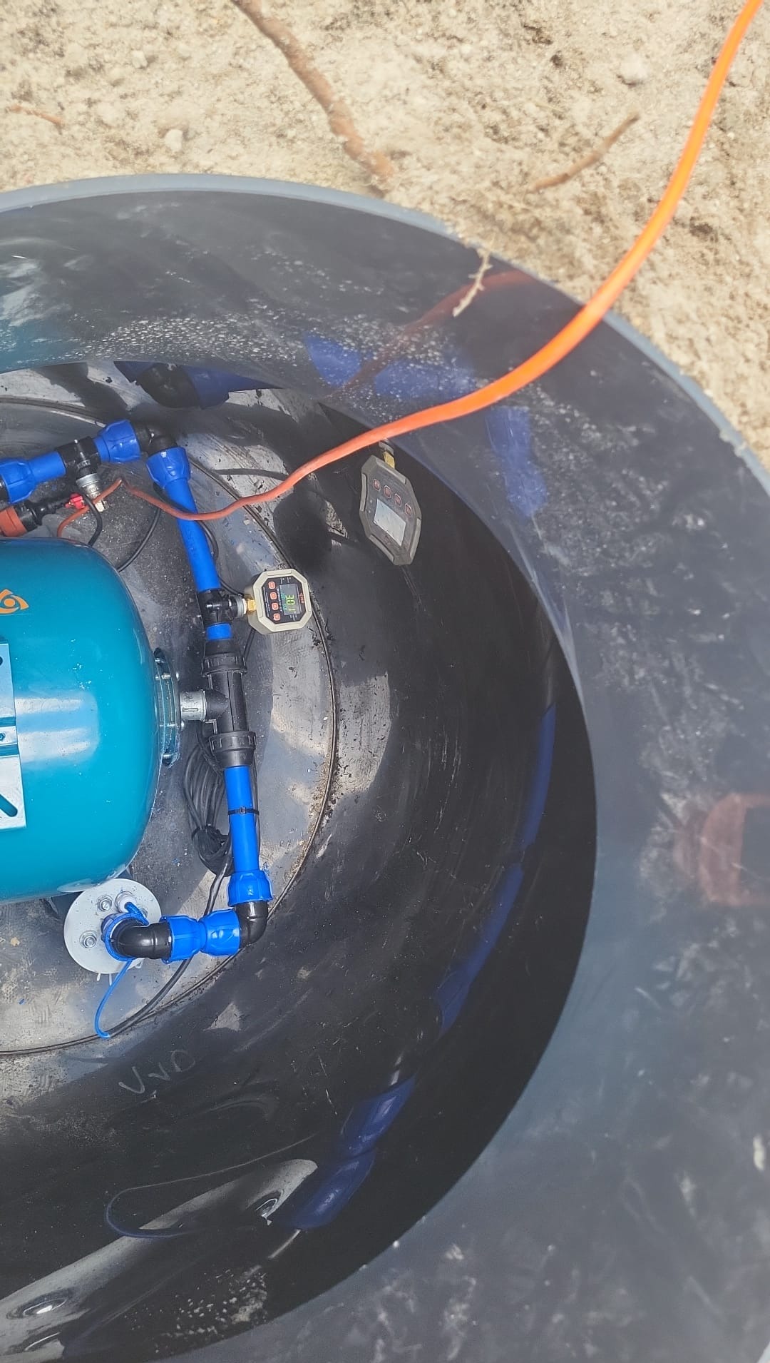 Wnętrze studni głębinowej z niebieskimi rurami, zbiornikiem hydroforowym i elektronicznym licznikiem. Widok z góry na instalację w czarnej obudowie.