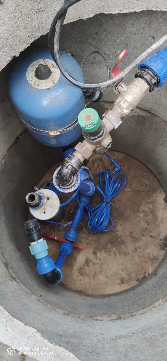 Wnętrze betonowej studni z niebieskim zbiornikiem hydroforowym, zaworami i przewodami. Widok z góry na instalację wodną wewnątrz studni.