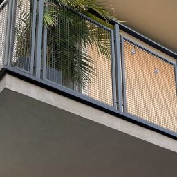 ROB TECH Robert Przewodowski - Metalowa balustrada balkonowa z siatkowym wypełnieniem i rośliną w donicy. Ujęcie z dołu ukazuje fragment budynku z widocznym balkonem.