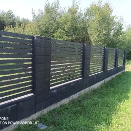 ROB TECH Robert Przewodowski - Nowoczesne ogrodzenie panelowe w kolorze grafitowym z betonowymi słupkami, otoczone zielenią. Widok na fragment domu i drzewa w tle.