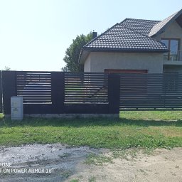 ROB TECH Robert Przewodowski - Nowoczesne ogrodzenie posesji z poziomych, ciemnych paneli, z widoczną skrzynką elektryczną i fragmentem domu w tle, w słoneczny dzień.