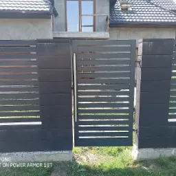 Nowoczesna, szara brama panelowa z poziomymi szczeblami, częściowo otwarta. W tle dom w stanie surowym i fragment ogrodzenia z elementami drewnianymi.