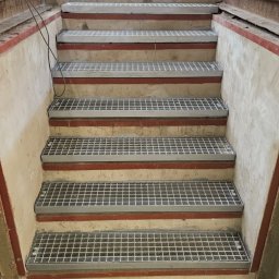 ROB TECH Robert Przewodowski - Metalowe schody kratowe w surowym wnętrzu hali przemysłowej, widok z dołu. Czerwone akcenty na stopniach, w tle widoczne światło dzienne.