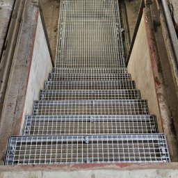 ROB TECH Robert Przewodowski - Metalowe schody kratowe w surowym otoczeniu przemysłowym, widziane z góry. Widoczne oświetlenie fluorescencyjne i fragment buta w dolnej części kadru.