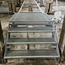 ROB TECH Robert Przewodowski - Metalowe schody kratowe prowadzące w dół, widok z dołu z perspektywą na tramwaj w tle. Konstrukcja stalowa w surowym, industrialnym otoczeniu.