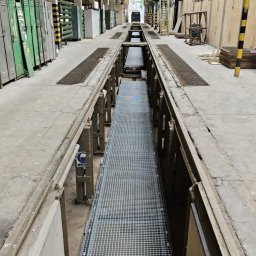 ROB TECH Robert Przewodowski - Długa, metalowa krata w kanale warsztatowym z widocznymi schodami wejściowymi, żółtymi suwnicami i rzędami szafek w tle hali.