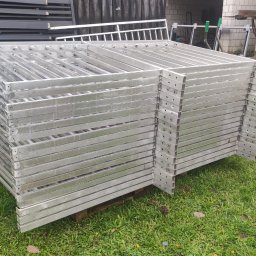 ROB TECH Robert Przewodowski - Stos srebrnych, metalowych ram ogrodzeniowych z otworami, ułożonych na palecie na trawie, z wózkiem widłowym w tle. Elementy gotowe do montażu.