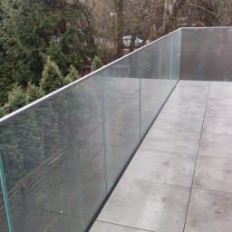 ROB TECH Robert Przewodowski - Szklana balustrada balkonowa z widokiem na drzewa. Nowoczesne rozwiązanie z płytkami podłogowymi w odcieniach szarości. Minimalistyczny design z elementami przyrody w tle.