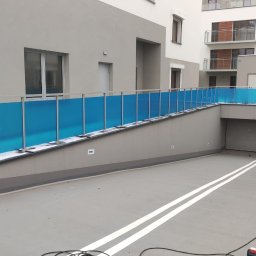 ROB TECH Robert Przewodowski - Nowoczesna balustrada ze szkła akrylowego w intensywnym odcieniu błękitu, zamontowana na rampie wjazdowej do garażu podziemnego w budynku wielorodzinnym.