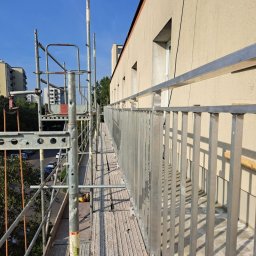 ROB TECH Robert Przewodowski - Montaż balustrady balkonowej z rusztowania. Widoczne elementy mocujące, konstrukcja budynku i fragmenty otoczenia miejskiego w tle.