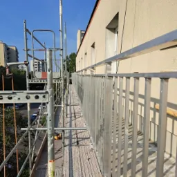 Montaż balustrady balkonowej z rusztowania. Widoczne elementy mocujące, konstrukcja budynku i fragmenty otoczenia miejskiego w tle.