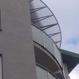 ROB TECH Robert Przewodowski - Nowoczesny balkon z zaokrągloną, szklaną balustradą i metalową konstrukcją zadaszenia, widziany z dołu na tle pochmurnego nieba.