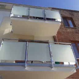 Dwa balkony z balustradami ze stali nierdzewnej i mlecznego szkła na tle budynku z drewnianą elewacją. Widok z dołu, słoneczny dzień.