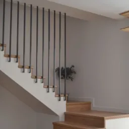 Schody drewniane z minimalistyczną, pionową balustradą w kolorze czarnym. Jasne ściany, drewniana podłoga. W tle widoczna roślina doniczkowa.