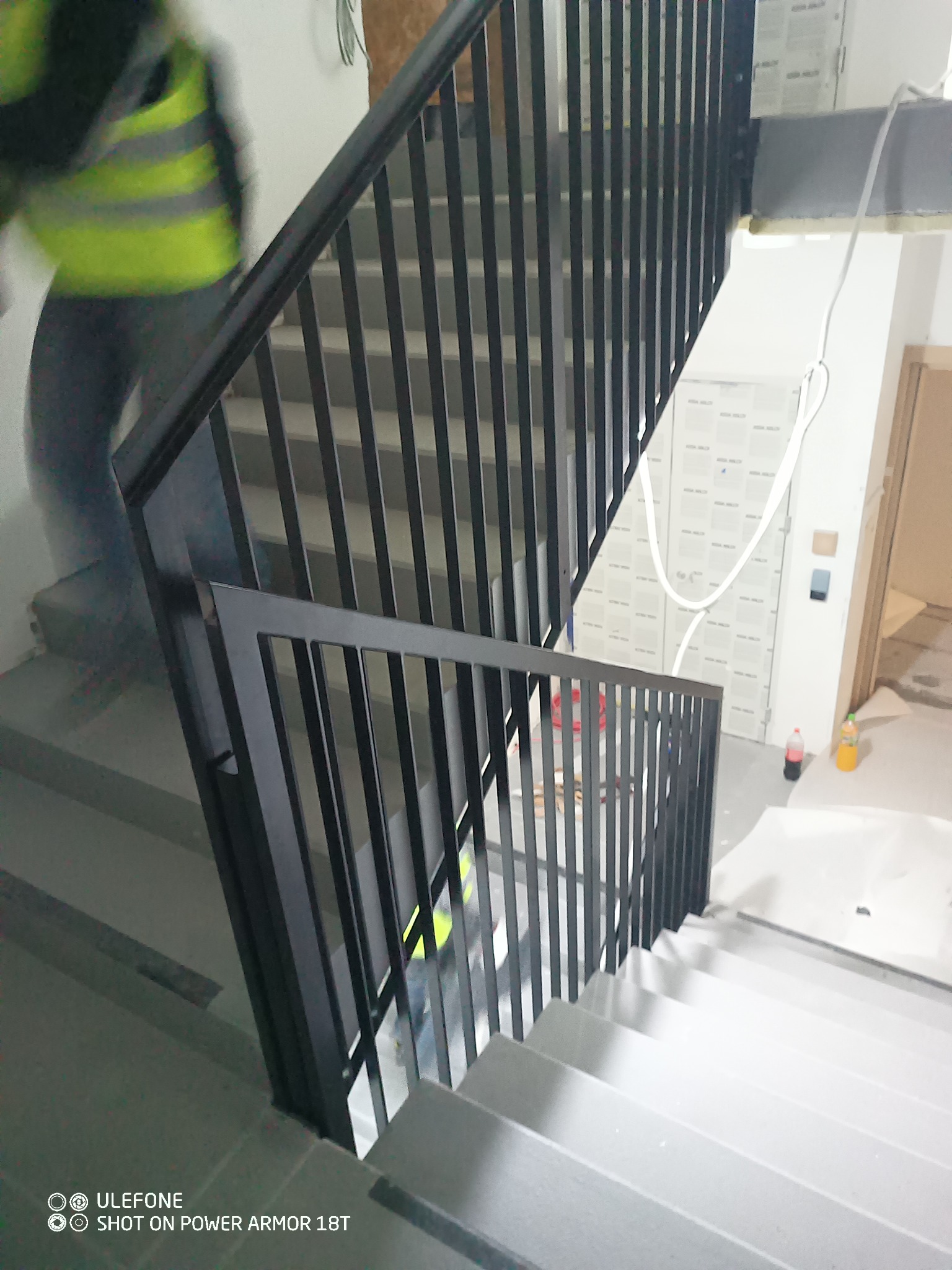 Nowoczesna, czarna balustrada metalowa na schodach wewnątrz budynku w trakcie wykańczania. W tle widoczny pracownik w kamizelce odblaskowej.