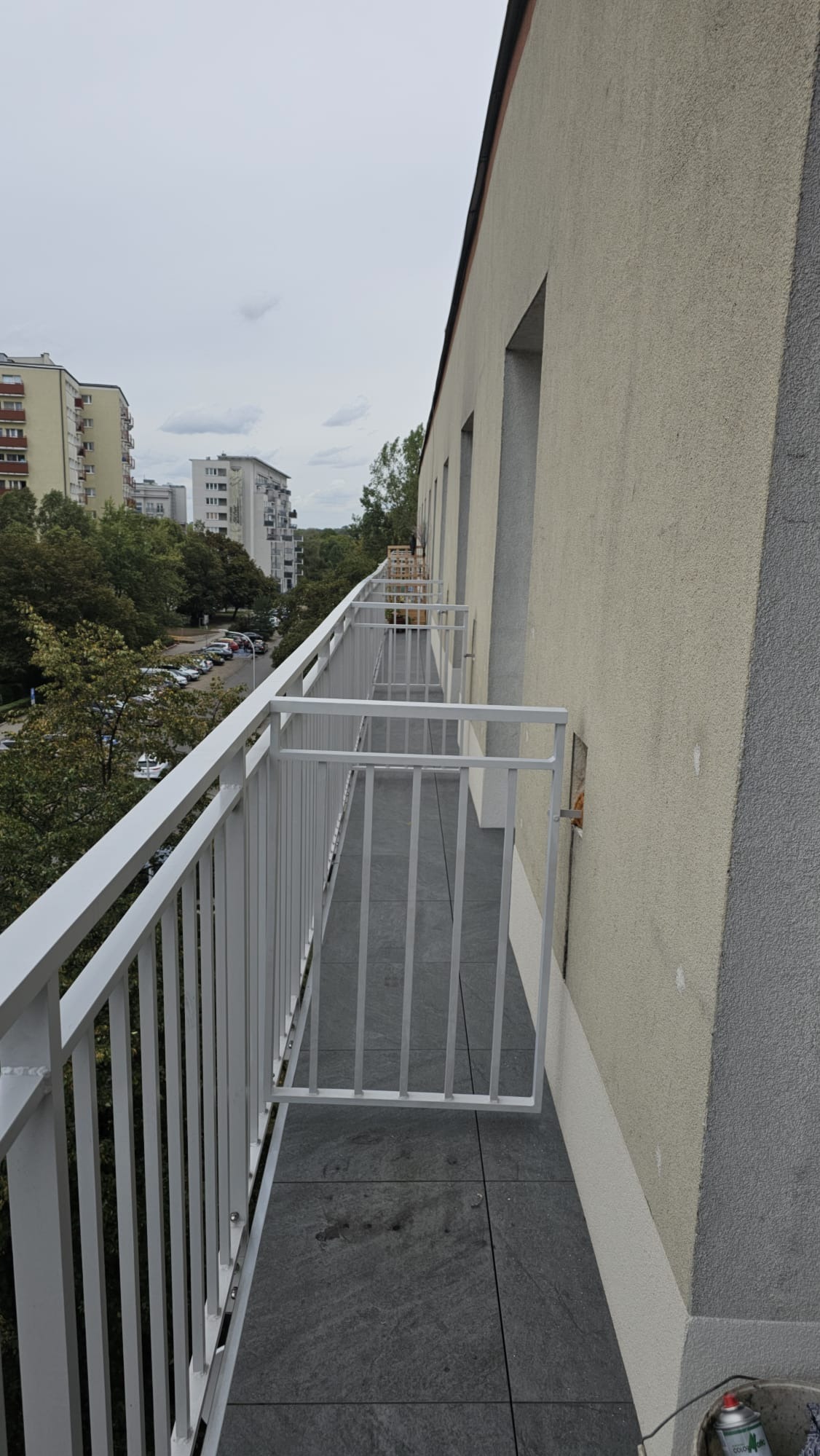 Biała balustrada balkonowa z furtką, widok z boku na tle zabudowy miejskiej i pochmurnego nieba. Szare płytki na podłodze balkonu.