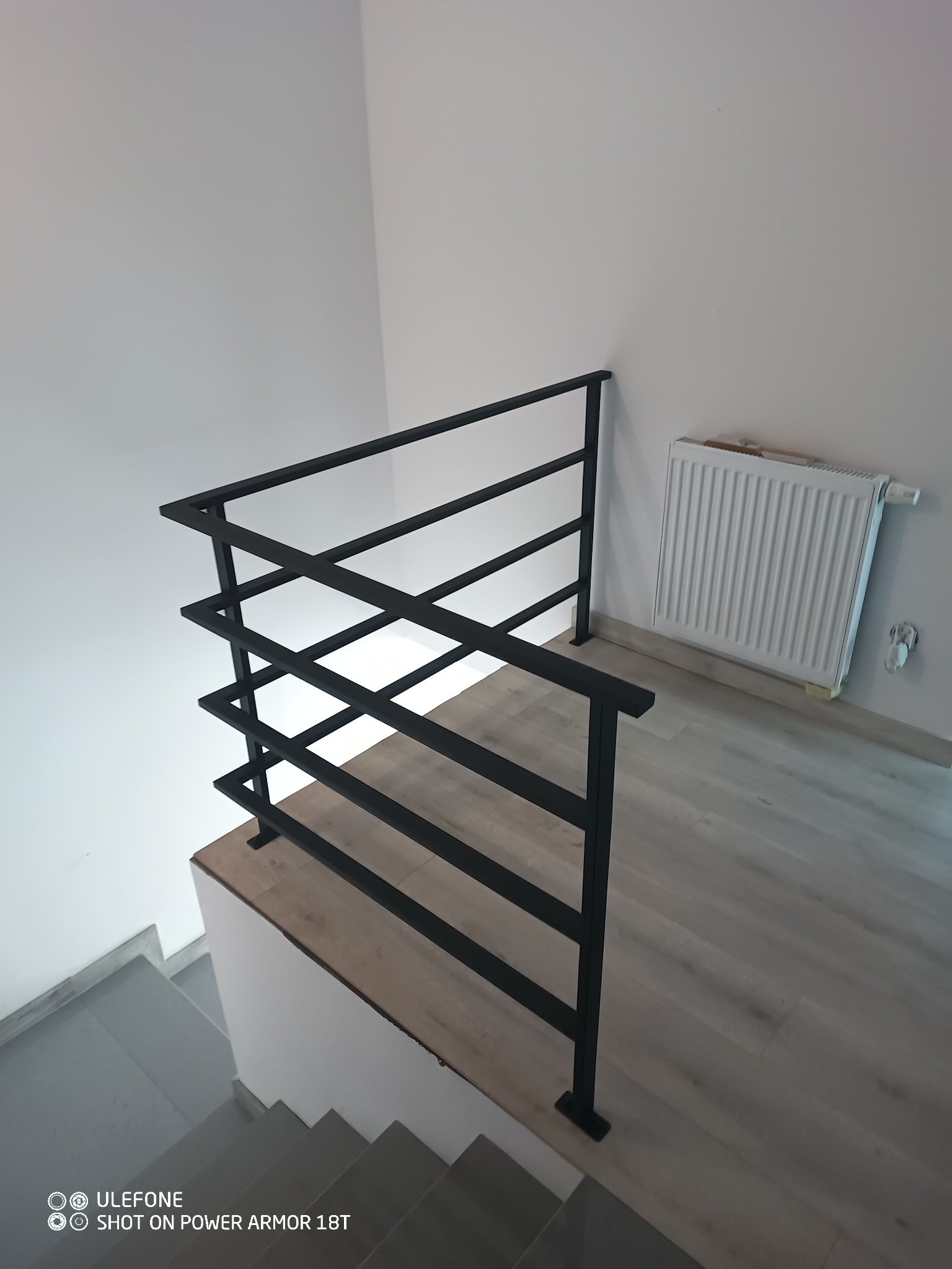 Nowoczesna, czarna poręcz schodowa z metalu, z prostymi, poziomymi szczeblami, zamontowana w jasnym wnętrzu, widok z góry na schody.